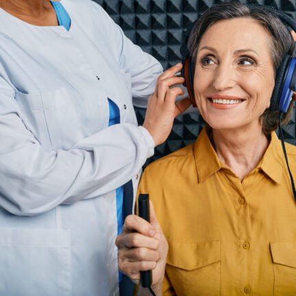 Portrait senior woman with white toothy smile while hearing check-up with ENT-doctor at soundproof audiometric booth using audiometry headphones and audiometer